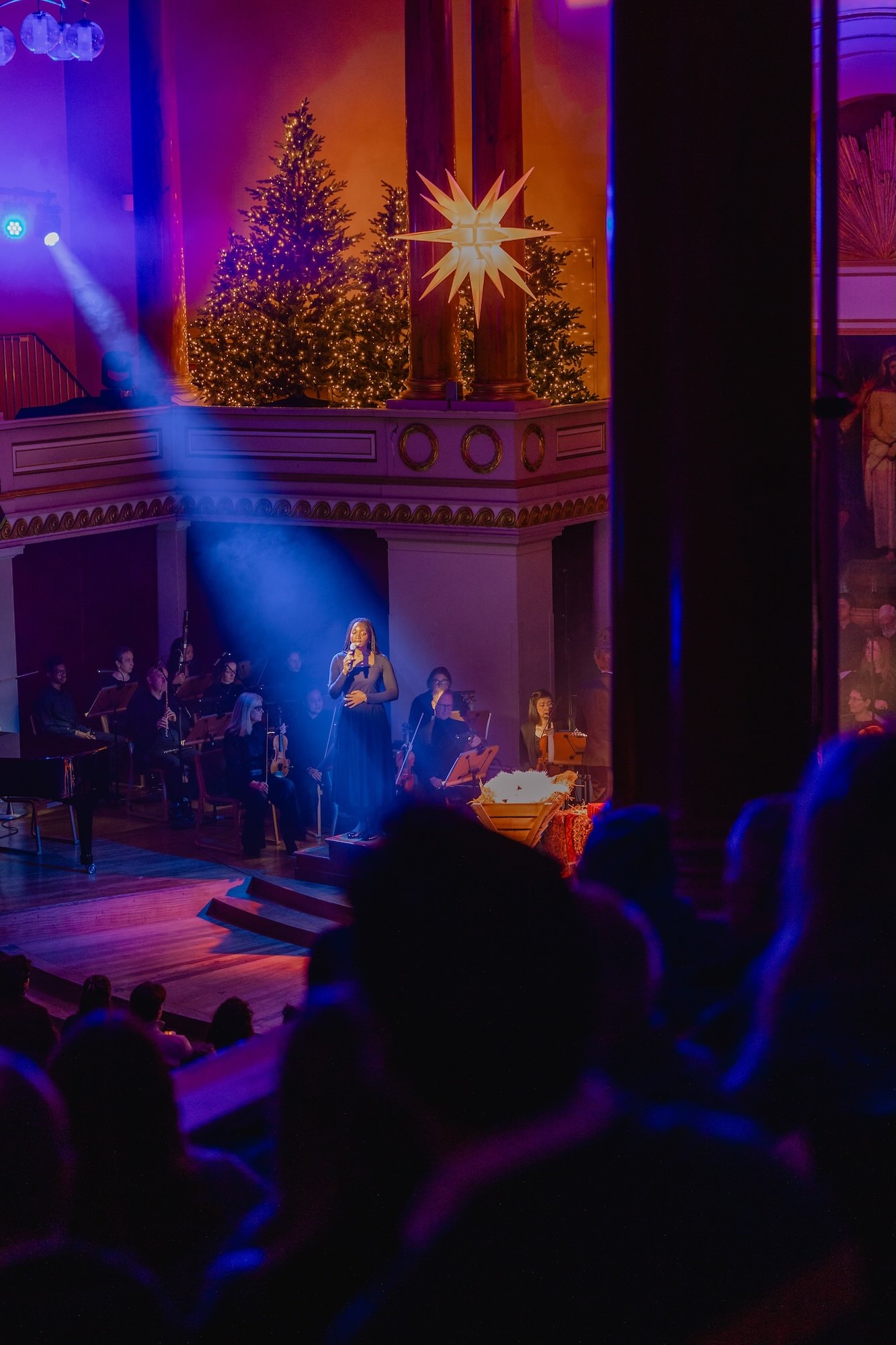 A soloist performs a carol at an All Souls carol service, framed by Christmas trees with festive lights, and a star hung above a manger full of hay.
