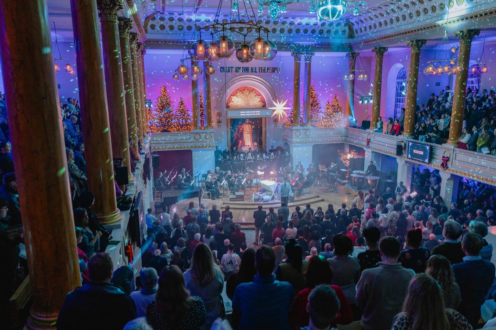 A Christmas carol service at All Souls. With an orchestra, singers, festive lights, manger, star and Christmas tree forest.